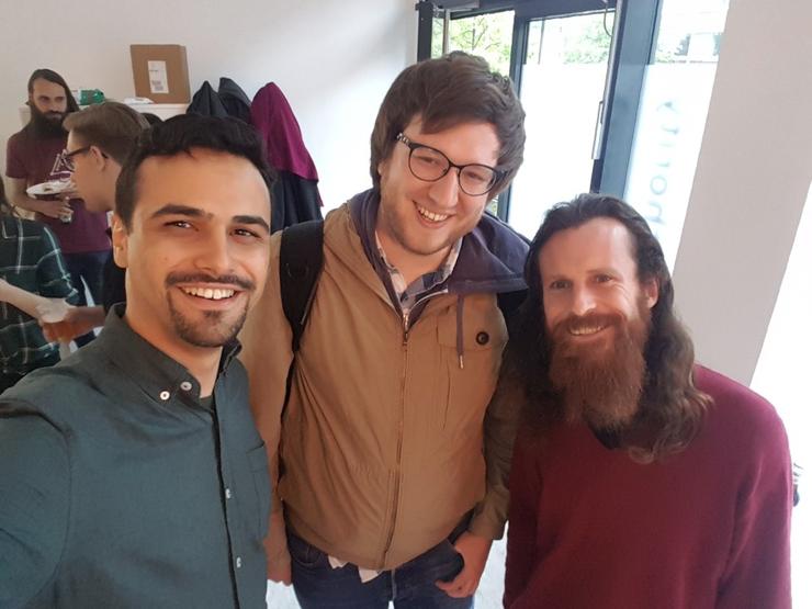 Photograph of Andre (left), Gordon (middle), and Dominic (right), looking towards the camera and smiling; they have long-sleeved shirts and are indoors, in a meetup event, where behind them there are a few people standing having conversations