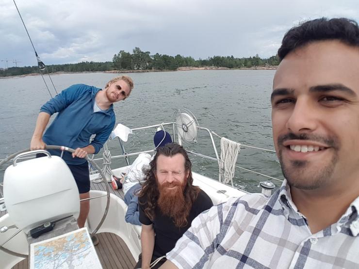 Photograph of (from left to right) Teemu Paivinen, Dominic Tarr, and Andre Staltz on a sailboat; on a partially clouded summer day, the sailboat is on the sea waters, with an island visible in the background; Teemu is steering the boat and making a tilted pose, Dominic is sitting down, and Andre is standing while taking the picture in selfie stance, all three are smiling