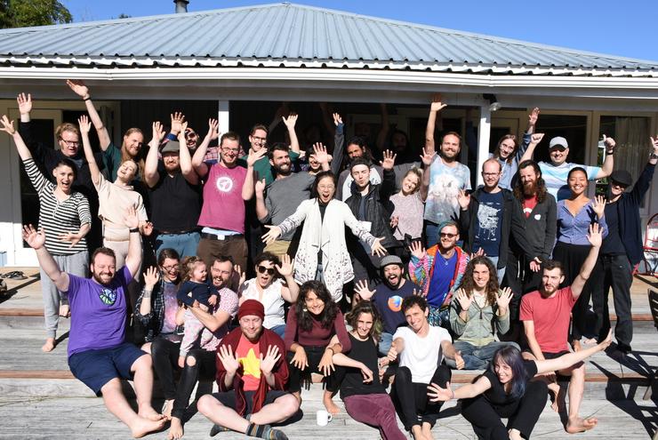 Photograph of approximately 40 people, some of them sitting on a wide wooden balcony staircase, some of them standing, and nearly all of them are raising their hands and smiling; they are outdoors, the sun is shining on them, and they are directly facing the camera; they are wearing comfortable clothes and many of them are barefoot